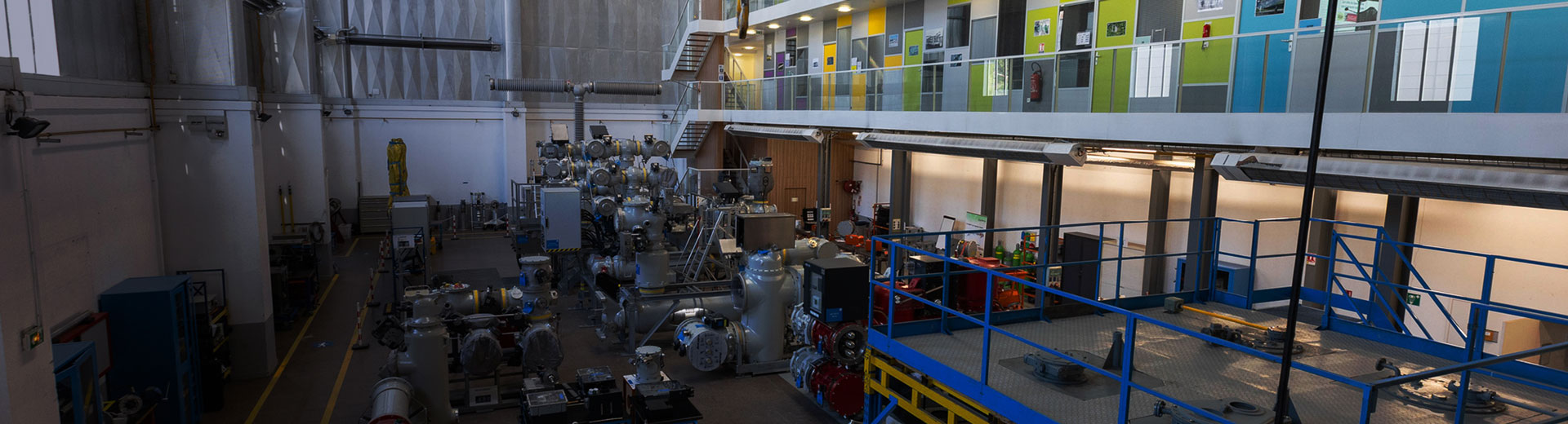 A wide, high-angle view of a technical training facility featuring industrial electrical switchgear equipment in the foreground and a multi-level office or classroom wing with colorful walls in the background.
